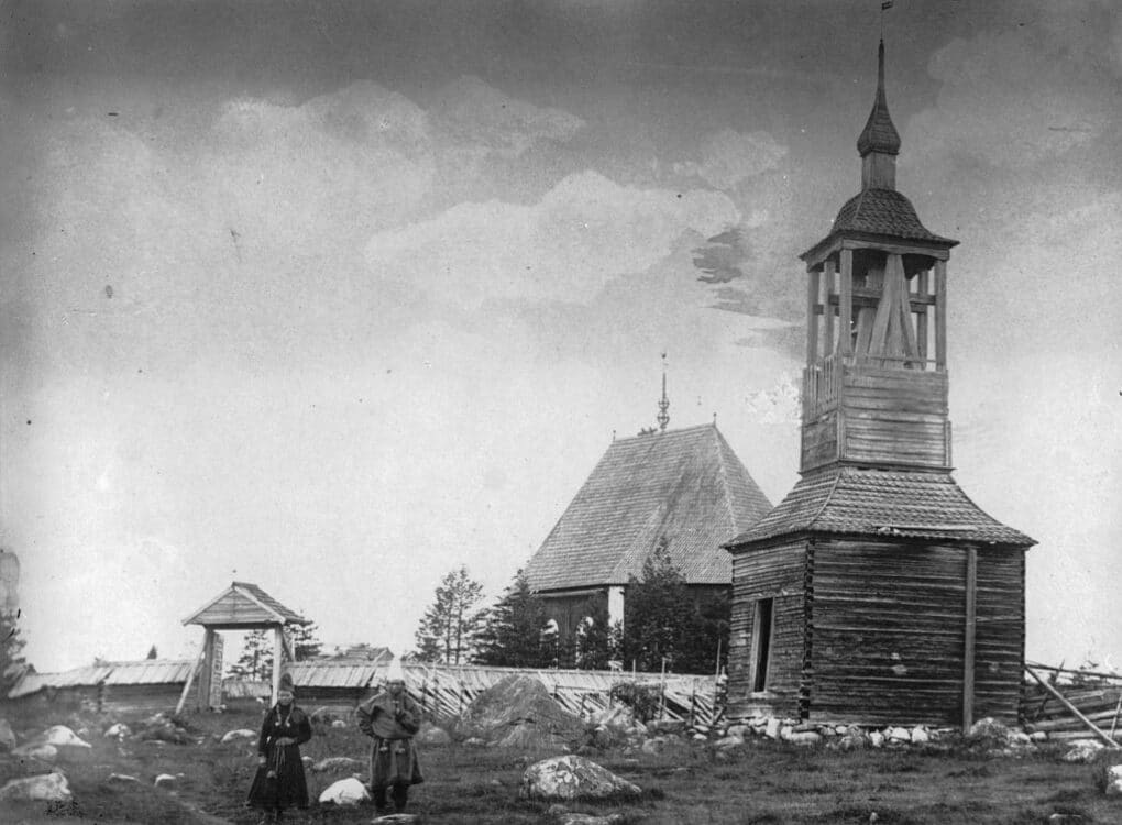 Black and white historic photo of very old wooden church with steeple; there is a couple in traditional Sámi dress standing in the foreground.
