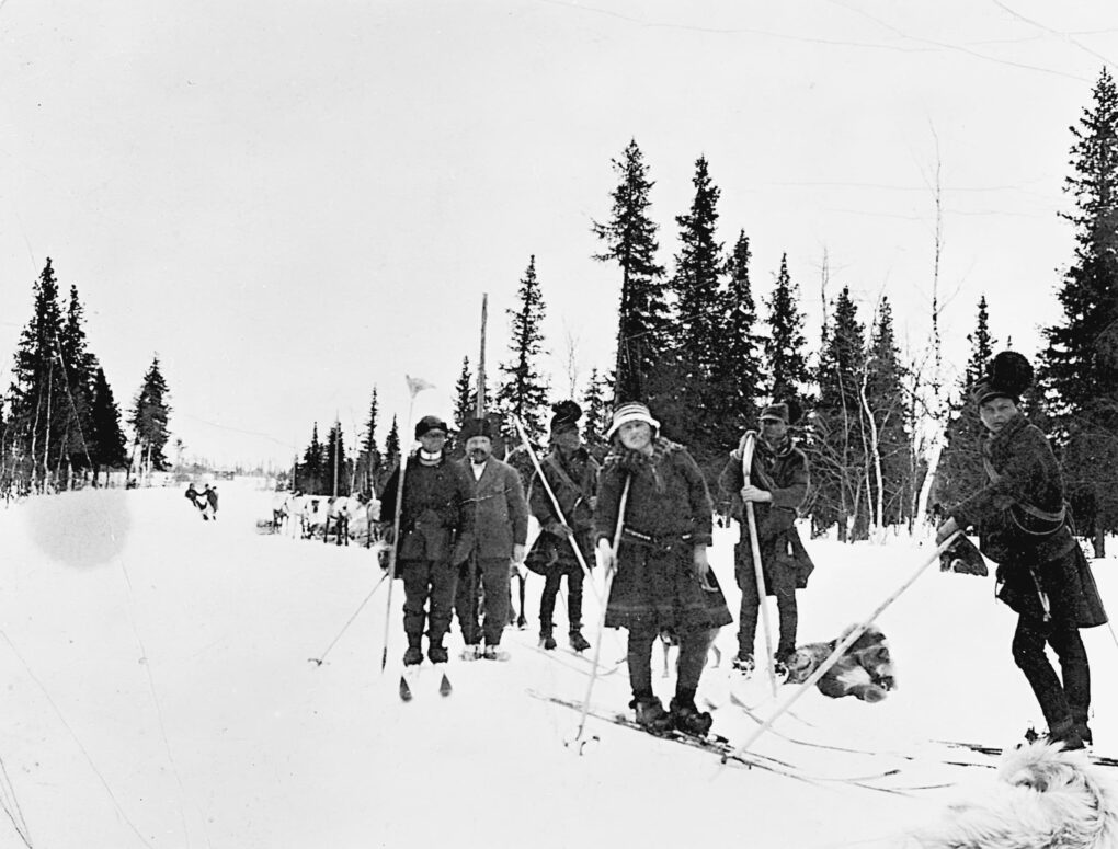 A procession of men on skis, paused and leaning on ski poles, in a snowy landscape with conifer trees.