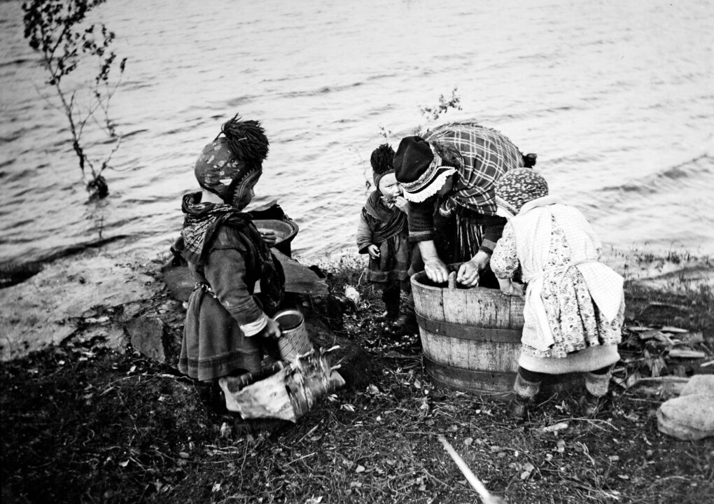 Black and white photo historical photo of woman bending over washtub on a gravelly shore. Several children helping her with laundry.