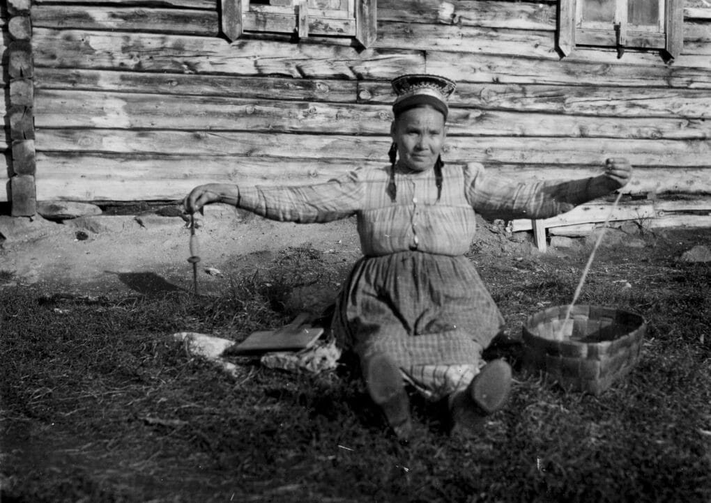 Black and white historical photo of woman seated on the earth with a spindle and a basket.