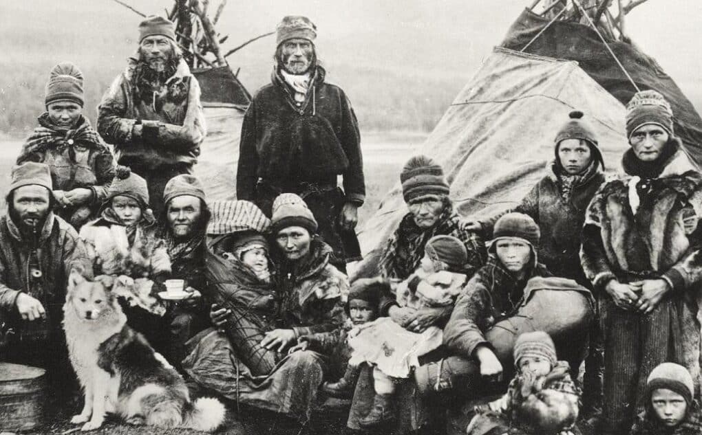 Black and white historical photo of group portrait of 16 Sámi, included several children and a dog, with teepees in background. 