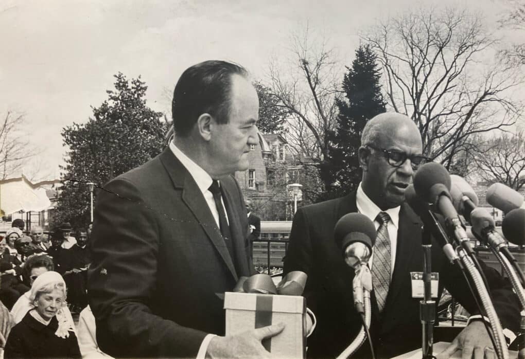 Black and white photograph of white politician and black business man at an outdoor microphone.