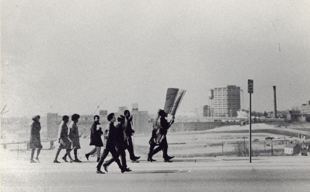 Black-and-white photo of a small group of black protestors carrying a sign, with a concrete office building in the distance.
