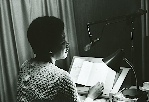 Click to Enlarge A woman with pearl earrings is reading from a book into a microphone. The book appears to be I Know Why the Caged Bird Sings, by Maya Angelou.
