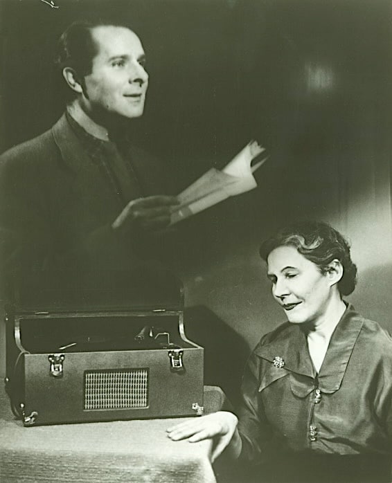 Click to Enlarge An older woman is listening to a record player, above which is a ghostly photo, superimposed, of a man reading.