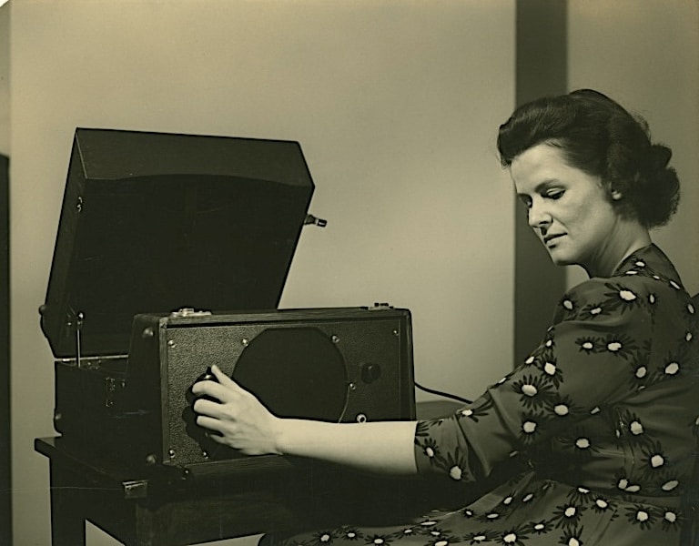 Click to Enlarge Sepia-tone photograph of a woman in a flowered dress adjusting the dial on an old-fashioned record player. She has her head tilted, and her eyes closed, as if intently listening.