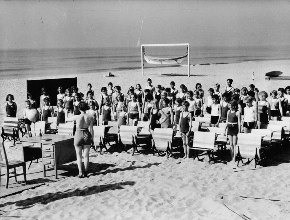 Photograph of school children in class on the beach, in sand, standing beside their small wooden desks. A woman teacher is giving instruction, also wearing a swimsuit, with a blackboard and desk. The ocean is visible not far in the distance.