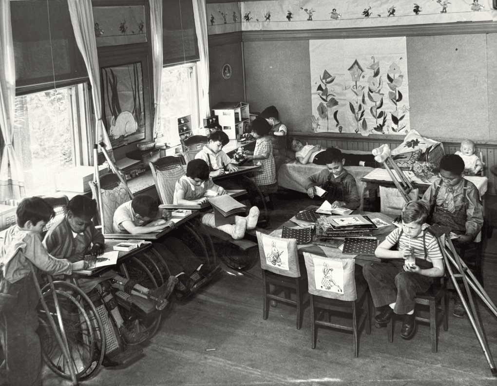 Eleven children seated in decorated classroom, using various mobility devices, and bent over their desk work. One child is lying down on a cot in the back of the room. Children are of various ethnicities, and appear to be between seven and ten years old.