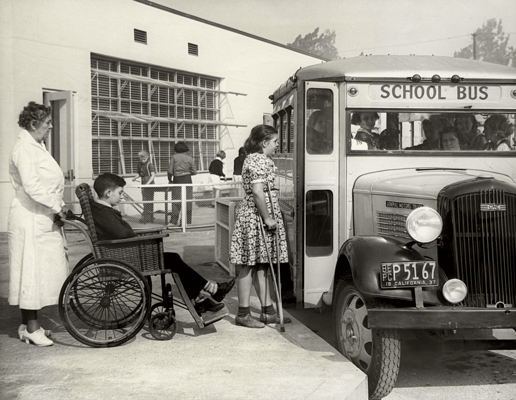 Two children waiting in line to board school bus, one girl using crutches, one boy seated in a wicker-weave wheelchair, being pushed by a nurse in a white uniform. Behind the children is a school with louvered windows.