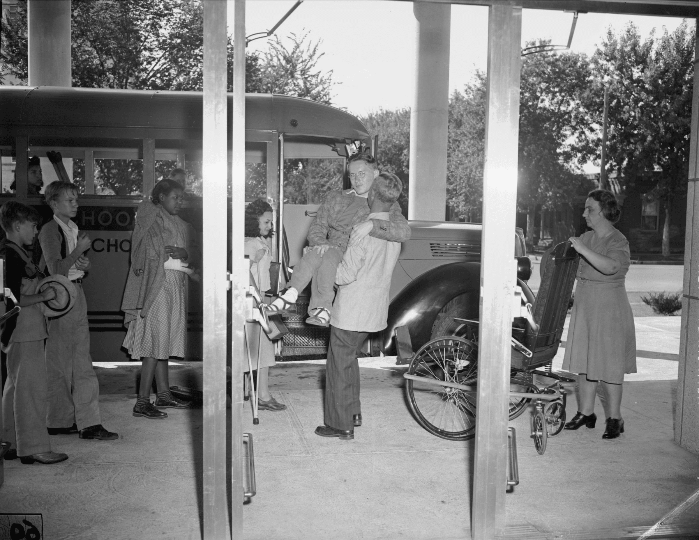 Click to Enlarge Outside near the school's front entrance, an aide or teacher is lifting a teenager from his wheelchair to the school bus while other students look on.