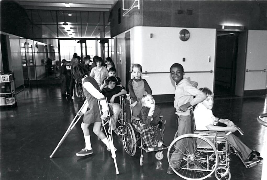 A dozen children seem to be standing in line inside the entrance of a school. There are several children in wheelchairs and several using crutches, and many are spontaneously smiling directly at the camera, as if photographed at play.
