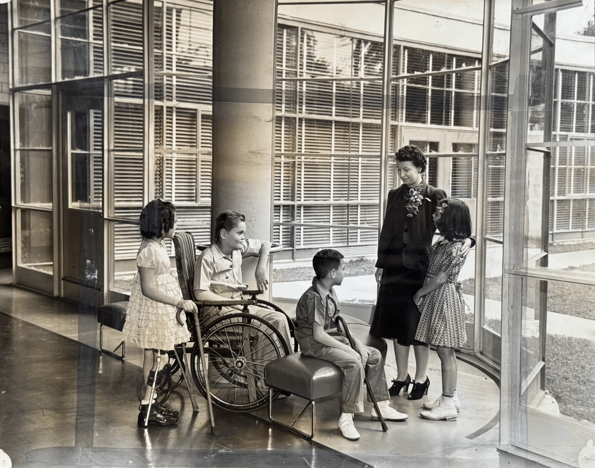 Click to Enlarge Obviously posed photo of smiling teacher with four students, two using a cane, and one seated in a wheelchair, standing in a rounded glass atrium space.