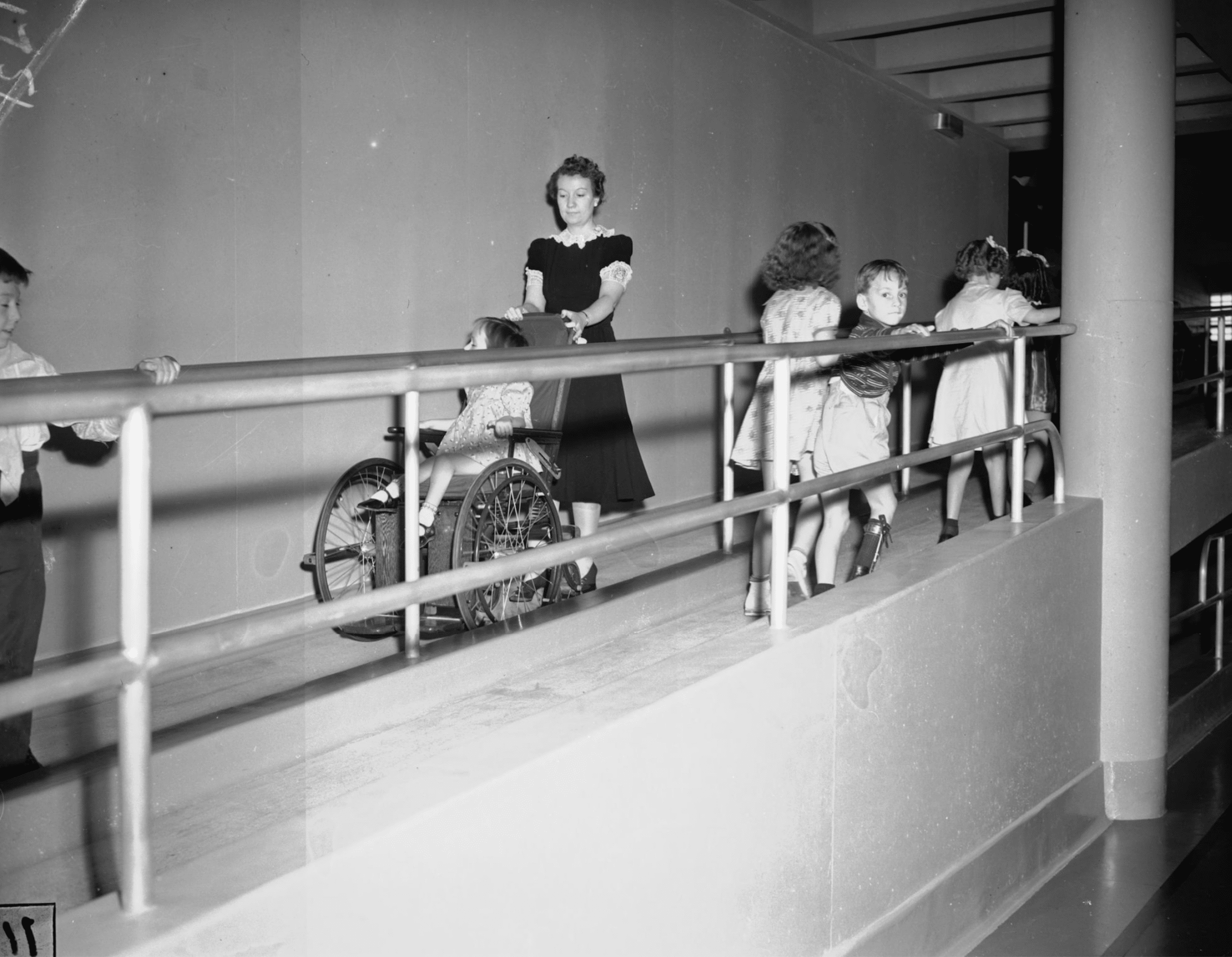 Click to Enlarge Five children are moving up the ramp, their hands on the railings, as a women is pushing another child down the ramp in a wheelchair. Children appear to be five to seven years old, and one boy wearing a leg brace is looking directly at the camera.