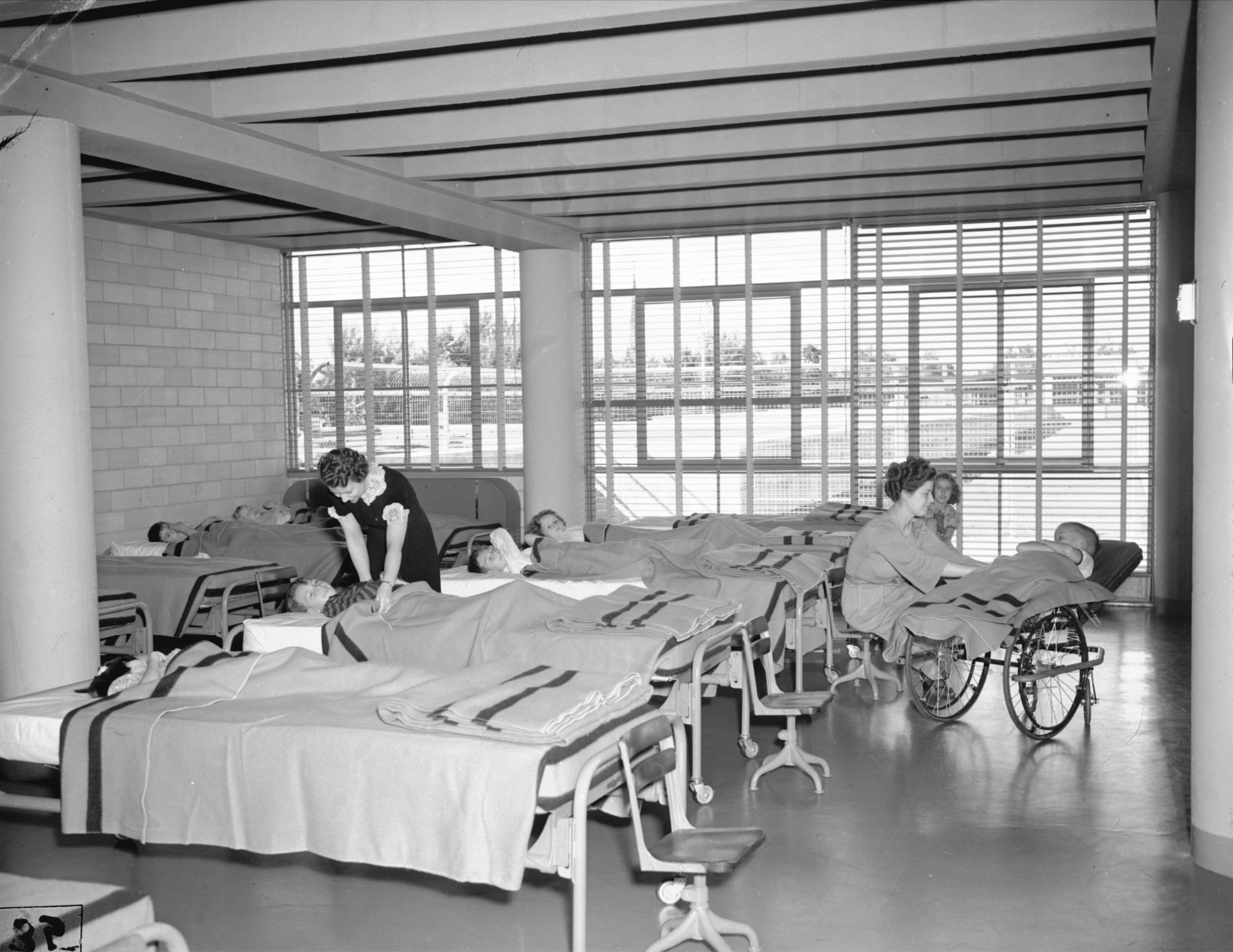 Click to Enlarge Approximately nine children are lying down, each in their own cot, with two women, presumably staff, tucking them in with wool blankets. The far wall is all windows and the blinds are open.