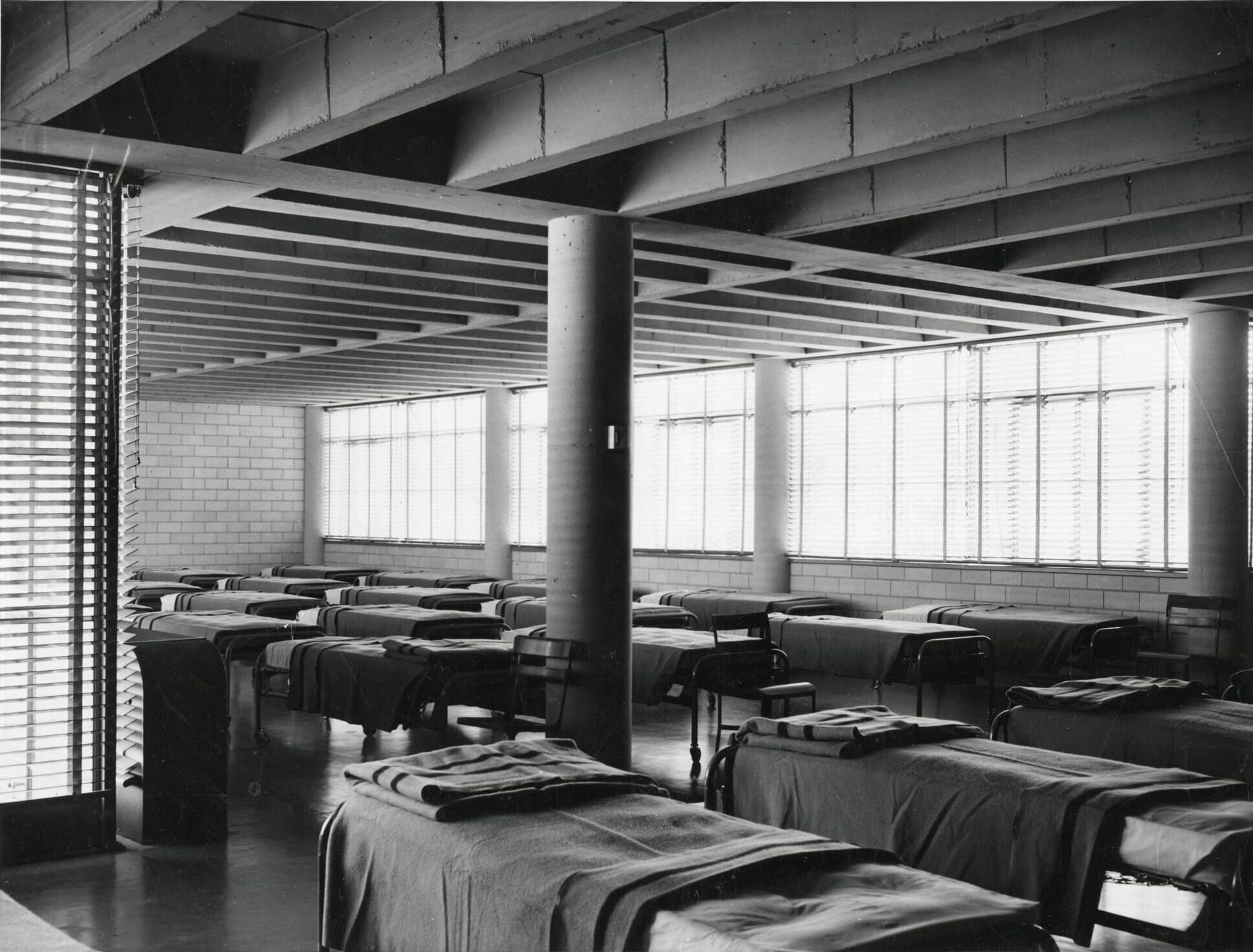 Click to Enlarge A large room with approximately twenty beds, with sheets and blankets. A large cement column is in the center of the room, with windows along the far wall. Photographic documentation of architecture — no students are present.