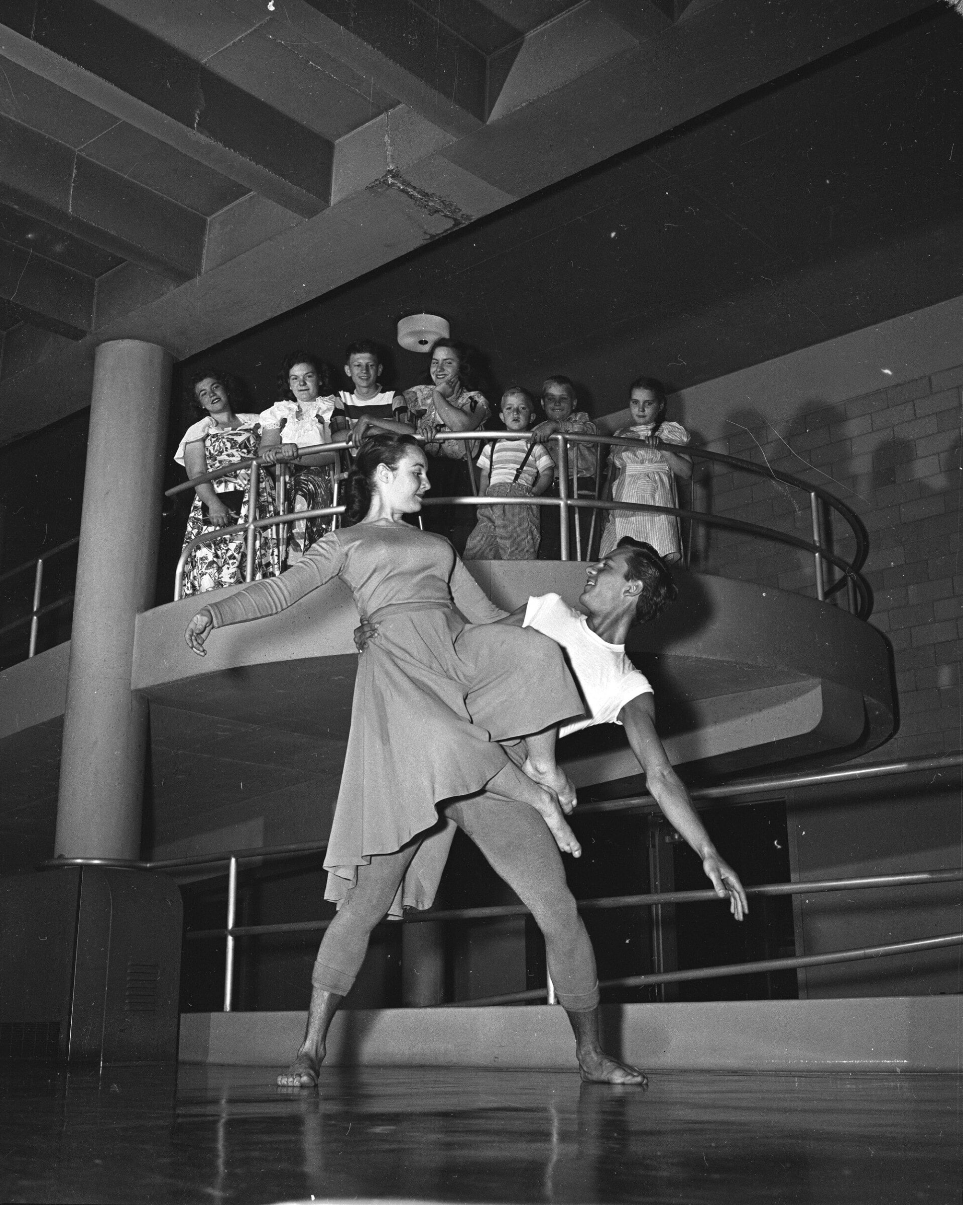 Click to Enlarge A group of students are standing on the second story of the school's interior ramp, watching a dance performance take place below them. A man and woman are dancing in bare feet; the woman is sitting on the man's hip, with her arm extended.