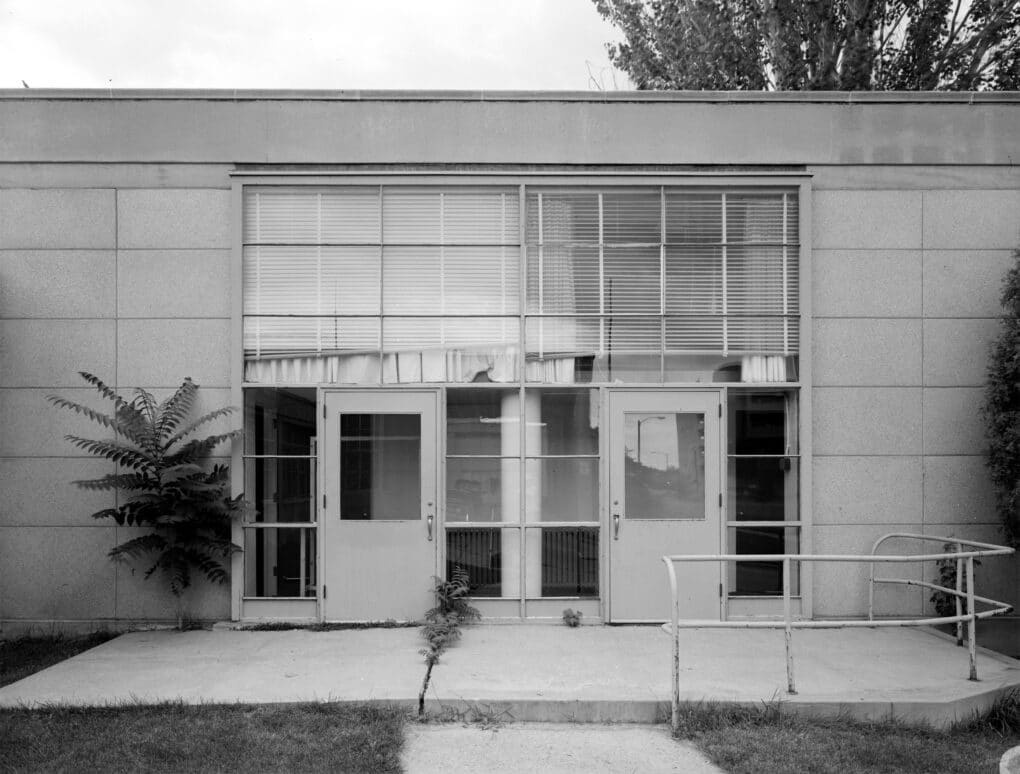 Side entrance to a school with two double doors. The concrete path has weeds and the window blinds appear to be broken.