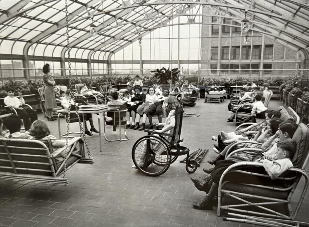 A large number of children are in a structure with class walls and ceilings that seems to have an expansive view. Some children are sharing swinging benches, some are in lawn chairs, and some are laying down. All three sides of the solarium are heavily planted with greenery. A teacher supervises.