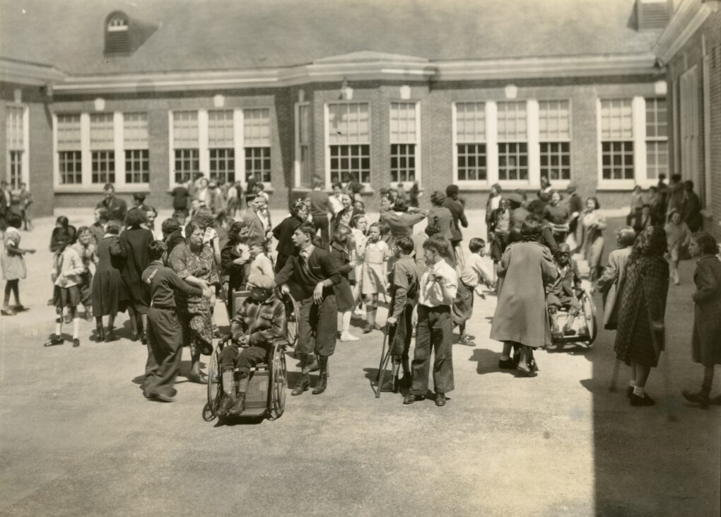 Black and white photograph of about forty children of all ages outdoors in an enclosed, concrete yard area, milling around and interacting with each other. There are many staff present as well, and many children have visible disabilities.