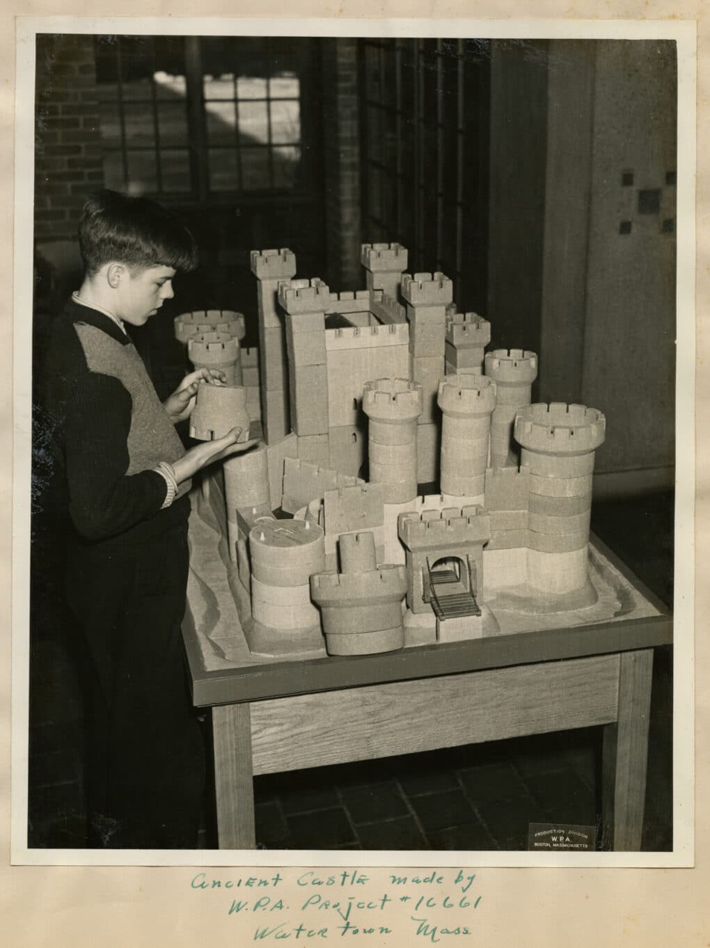 Photograph, in profile, of a Perkins Upper School student exploring a model of a castle. The light-skinned student, who is wearing a sweater over a shirt and tie, is holding a removable piece of a tower. The model includes several towers and a surrounding moat. The door is lowered over the moat, suspended by chains.