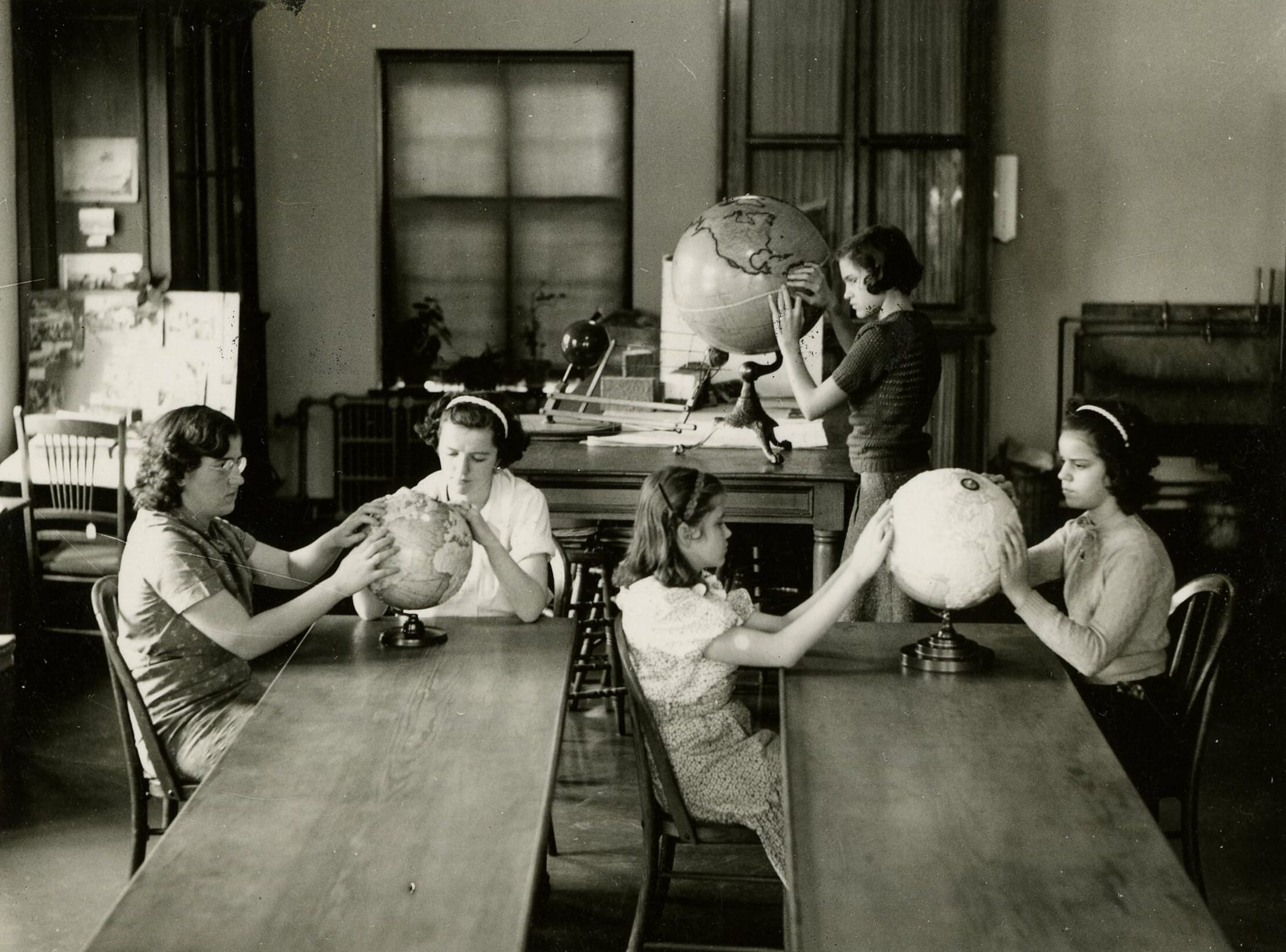 Click to Enlarge Five young women, aged approximately 11 to 14, explore three tactile globes on long wooden tables.