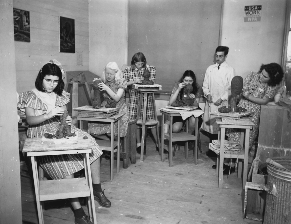 Five young women are intently sculpting what appear to be busts out of clay in a small art studio. A male teacher wearing a white smock assists.