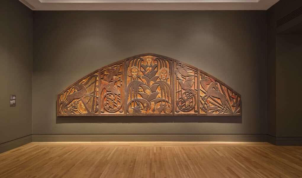 Large, heavy, finely carved wood five-panel sculpture hangs flush against the wall, with faces, animals, and music notes in various colors of varnished wood.