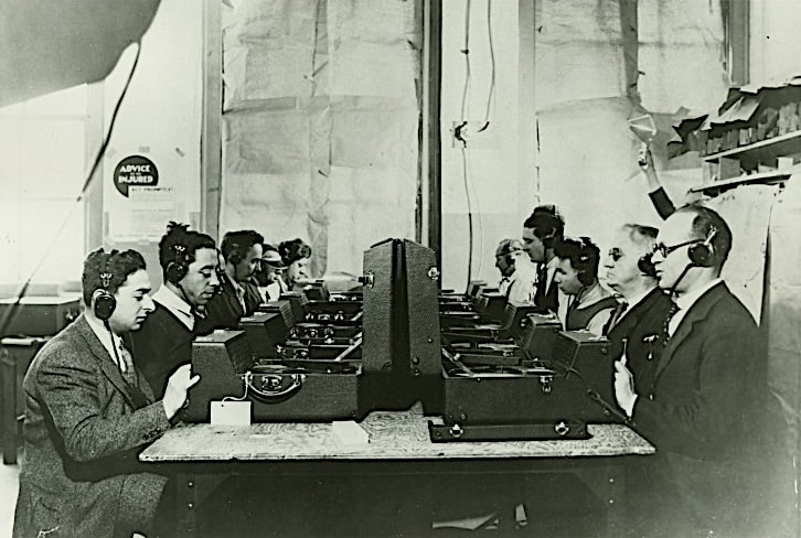 Click to Enlarge Black and white photograph of a long table, with about ten people seated, wearing headphones, with a boxed stereo in front of them, presumably listening and testing audio of recorded books.