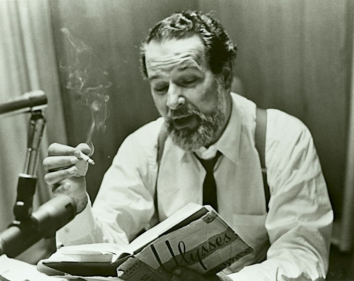 Click to Enlarge A middle-aged man is reading an open book into a microphone. He is also smoking. The book is Ulysses by James Joyce.
