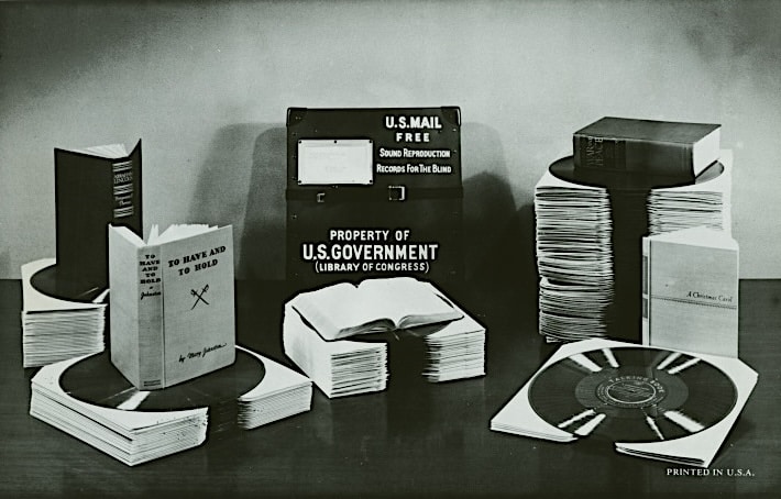 Click to Enlarge Black and white staged photo of several open books, including A Christmas Carol, sitting atop stacks of records. In the center is a metal box on which is printed,