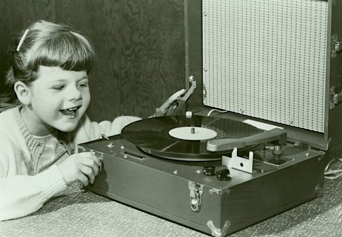 Click to Enlarge A young girl of about five years old is excitedly watching and listening to a record. She is holding and controlling the record player knobs.