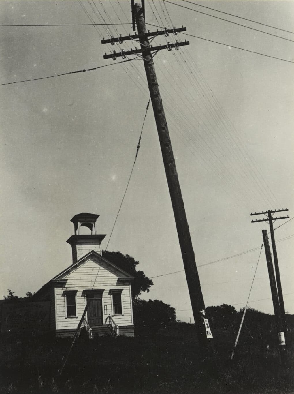 Black-and-white photograph of white clapboard school with bell tower; in the foreground are looming electrical poles and wires.