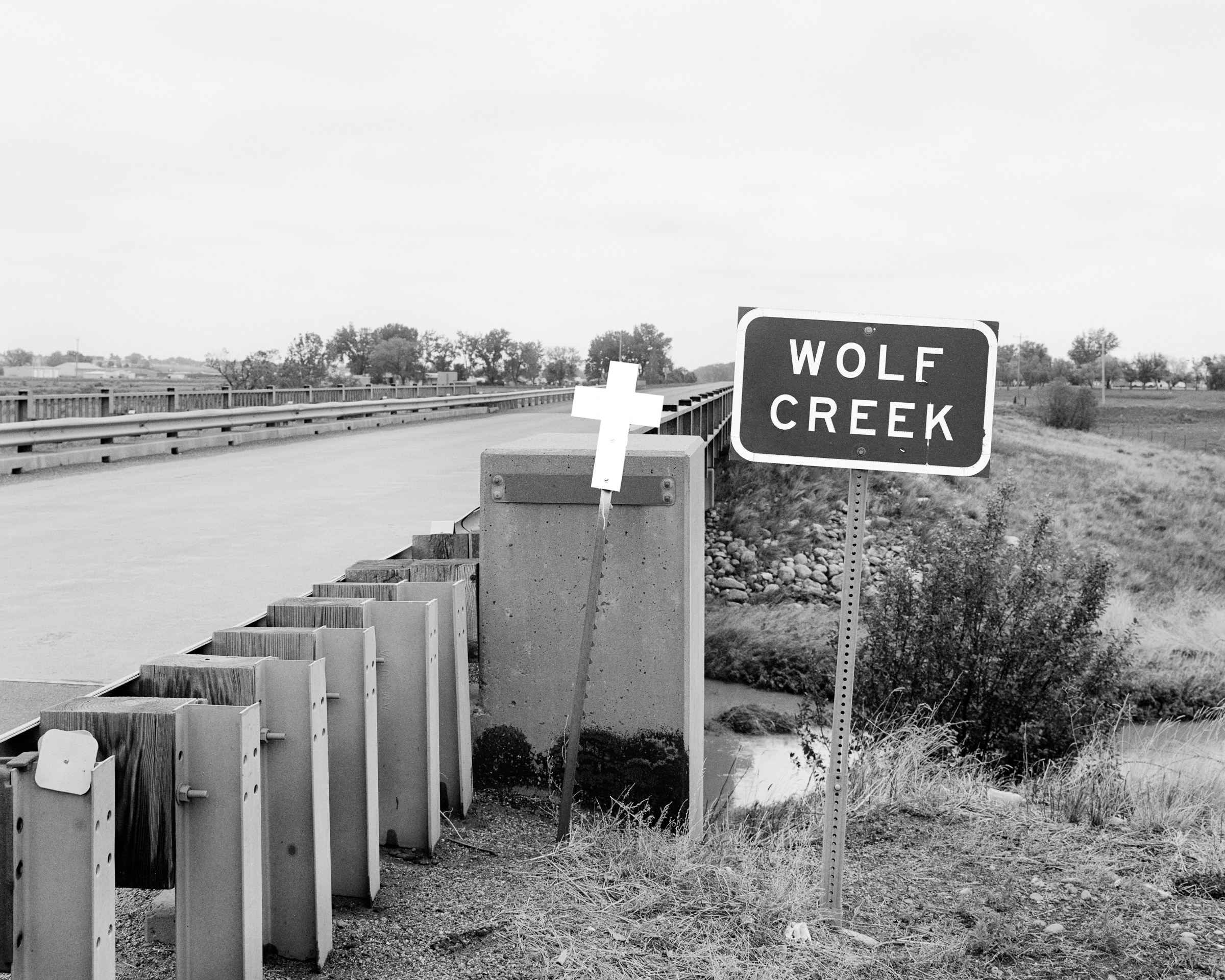 Click to Enlarge Black-and-white photograph of small white cross on metal pole beside highway in a flat, rural landscape, beside a small stream and a sign reading WOLF CREEK.