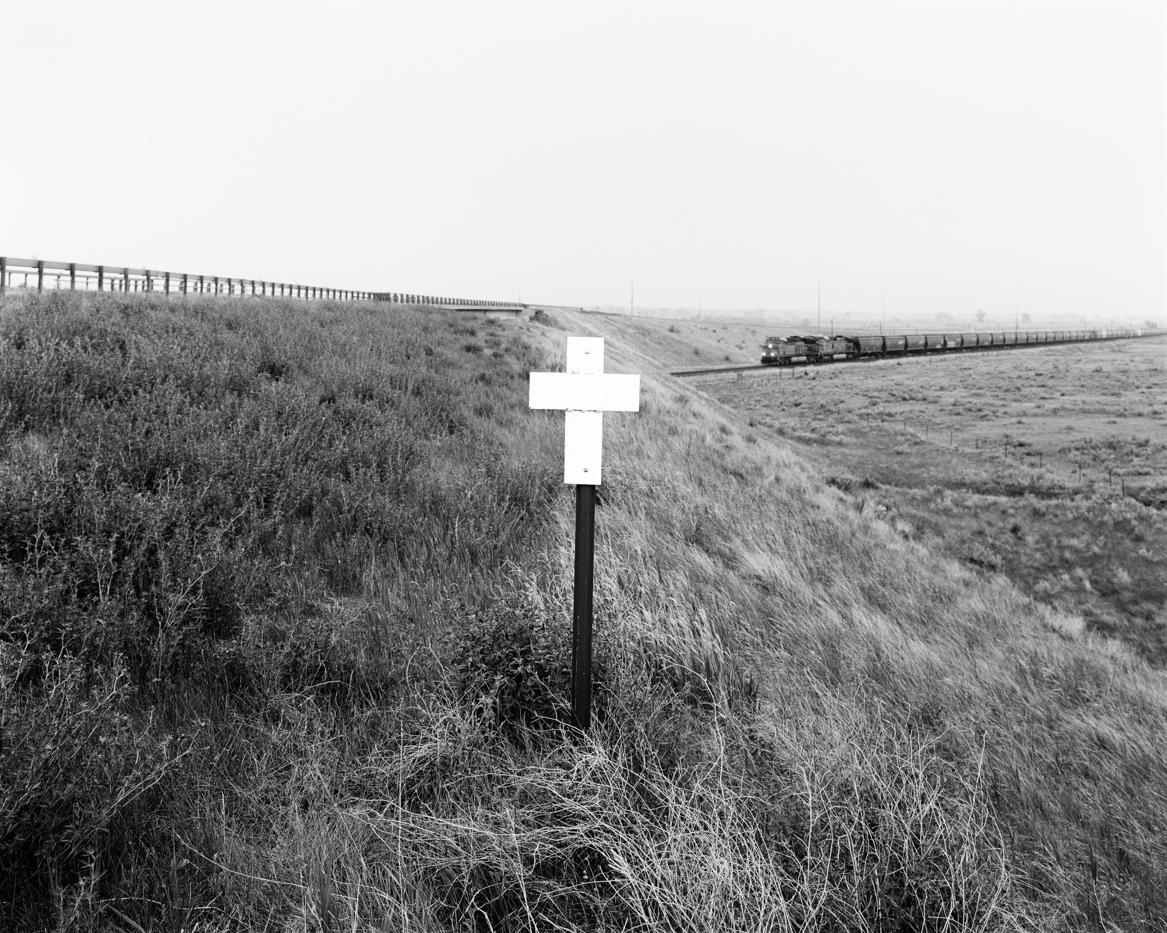 Click to Enlarge Black-and-white photograph of white cross on a pole on grassy slope below highway barrier, with a train in the midground.