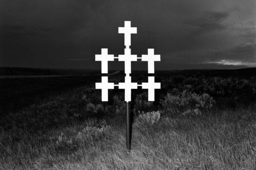 Black-and-white photograph of seven white crosses, mounted on a pole, in a dark field of grass.