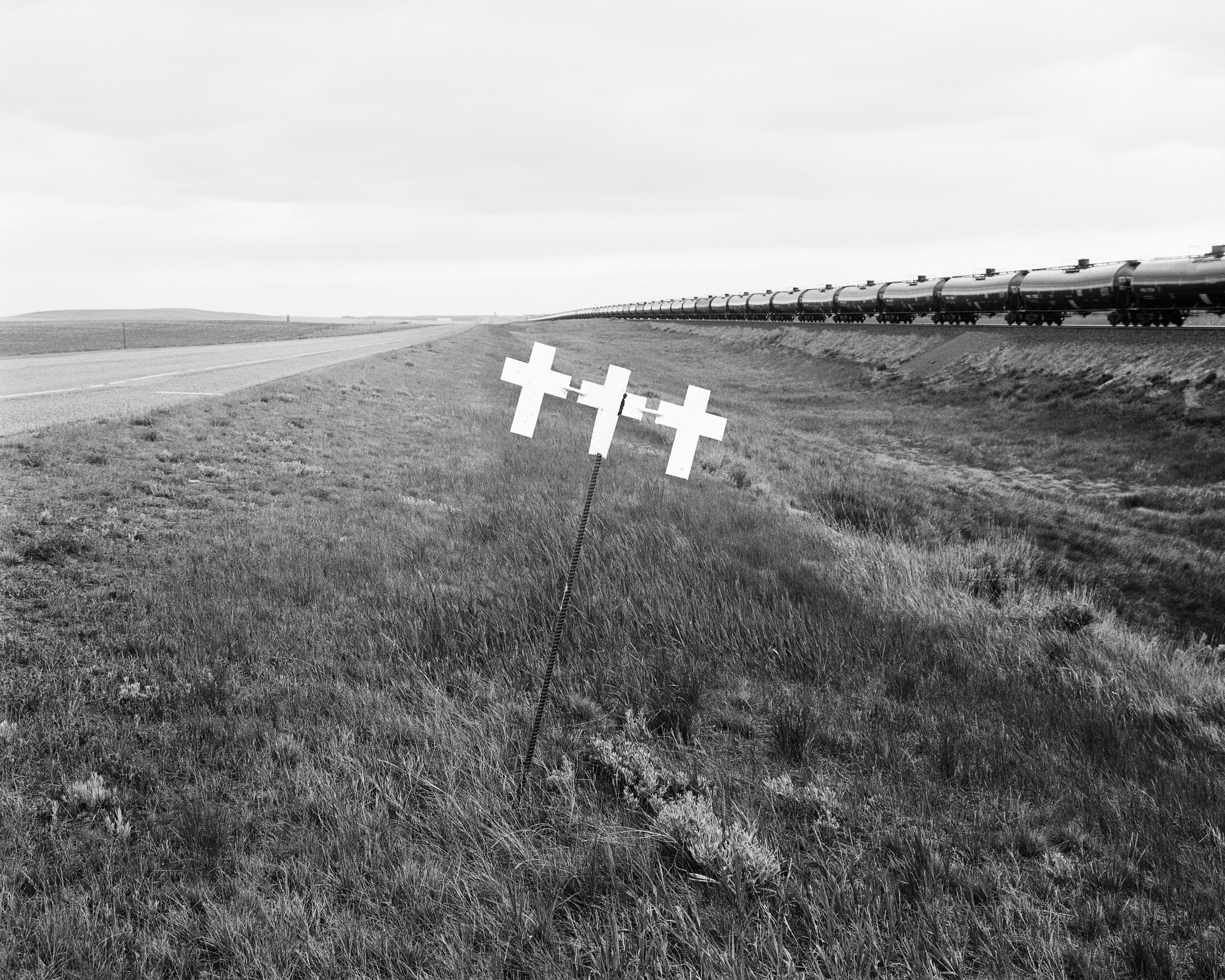 Click to Enlarge Black-and-white photograph of three white crosses on a grassy slope beside a road, with a train running through the midground from the right.