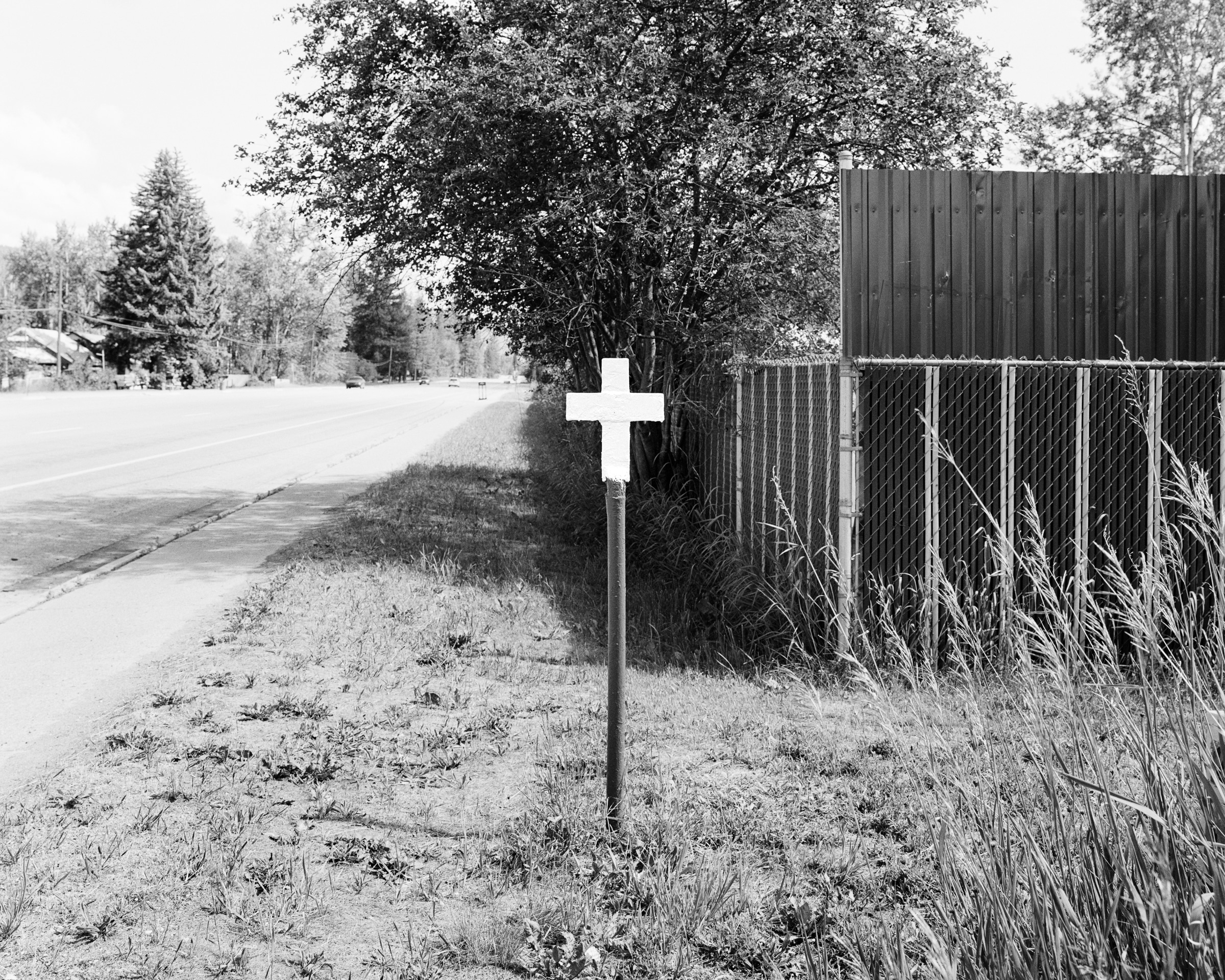 Click to Enlarge Black-and-white photograph of white cross on a pole beside a small-town road and chainlink fence.
