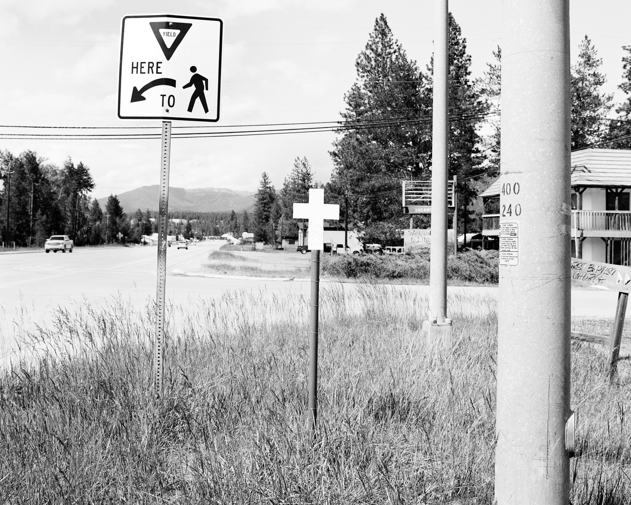 Click to Enlarge Black-and-white photograph of white cross on a pole at a small-town intersection.