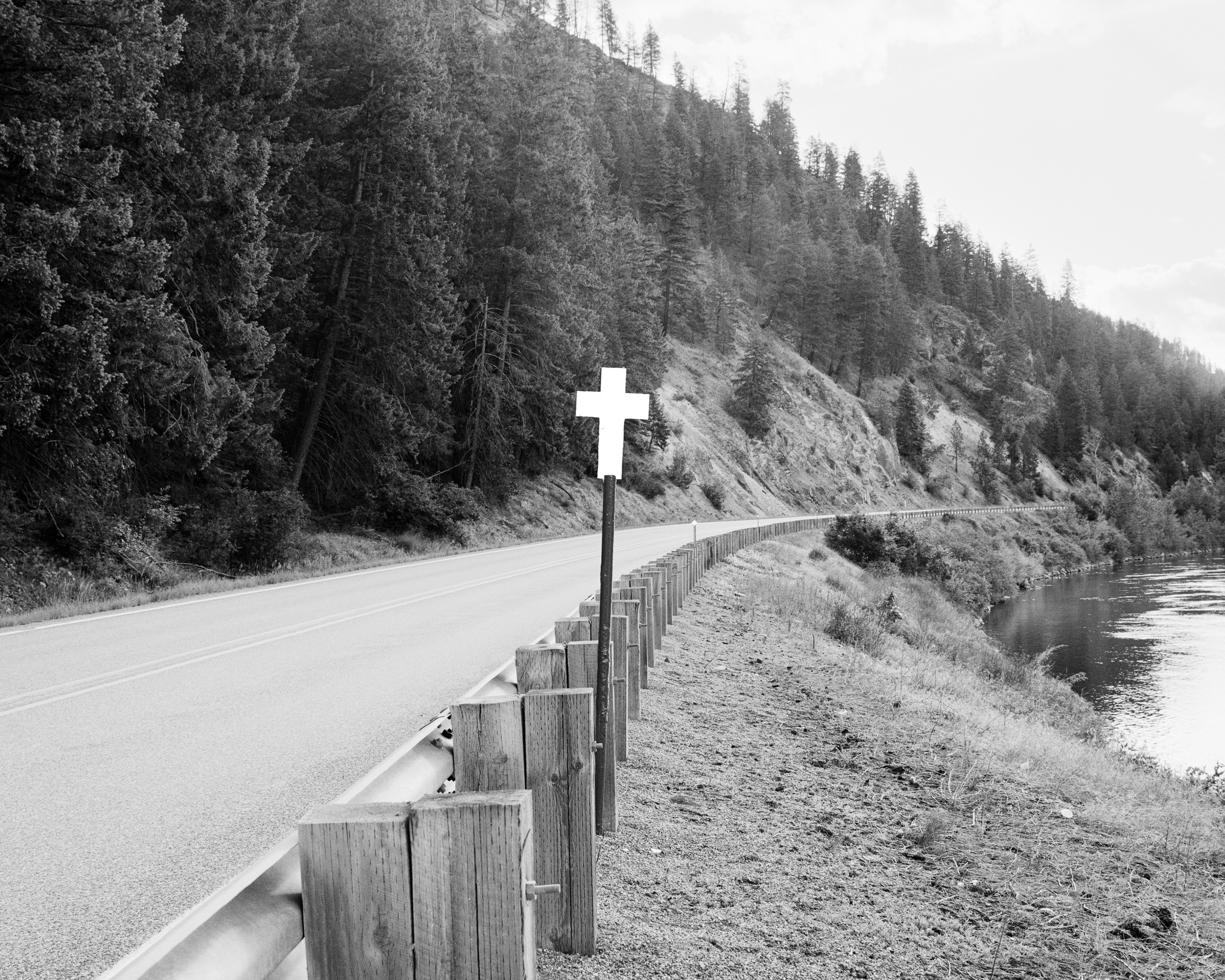 Click to Enlarge Black-and-white photograph of white cross on a pole beside a rural highway with a wooded hill at left and a lake or river at right.