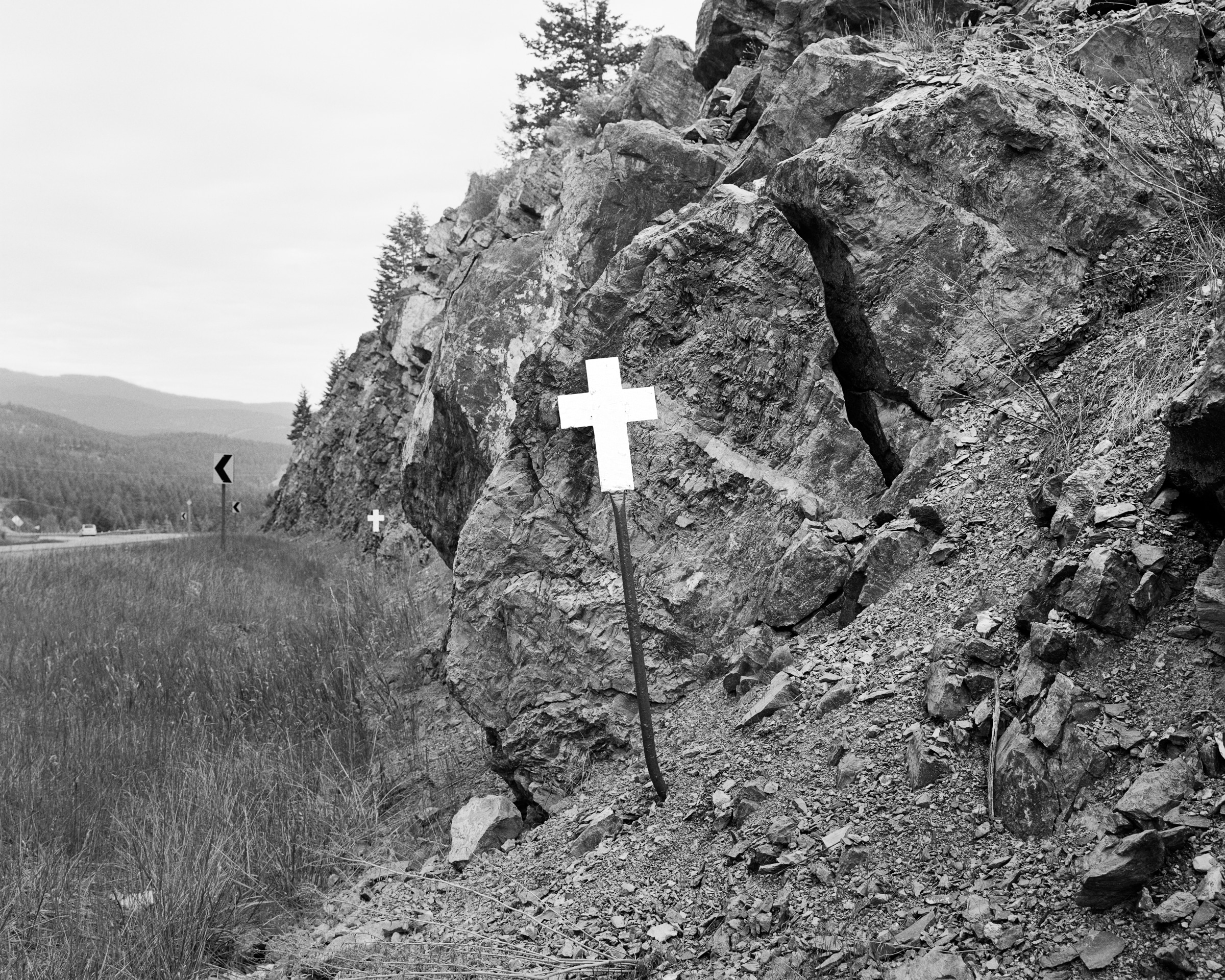 Click to Enlarge Black-and-white photograph of white cross on a pole beside a rocky outcrop, with highway at far left.