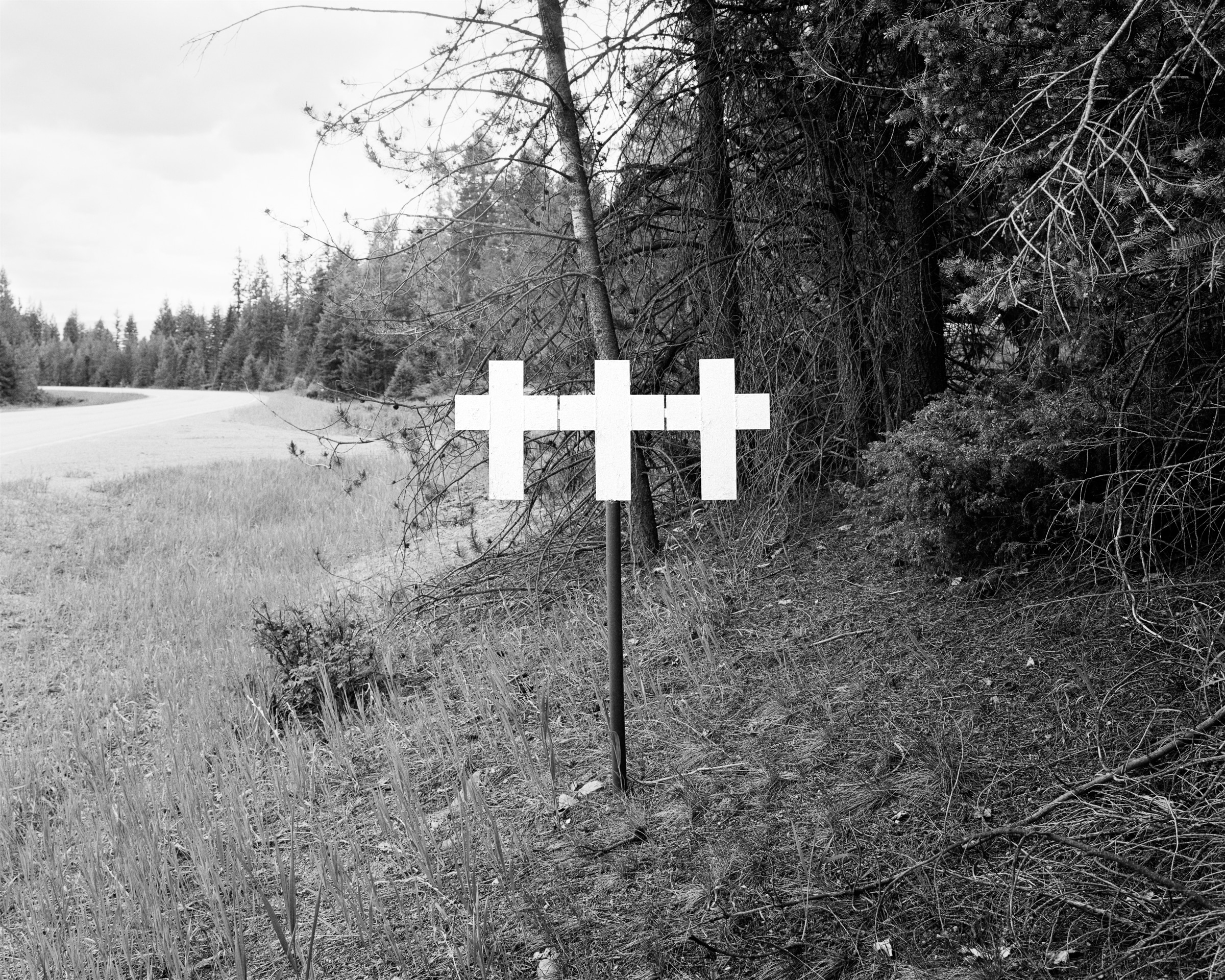 Click to Enlarge Black-and-white photograph of three white crosses on a pole on wooded alongside road.