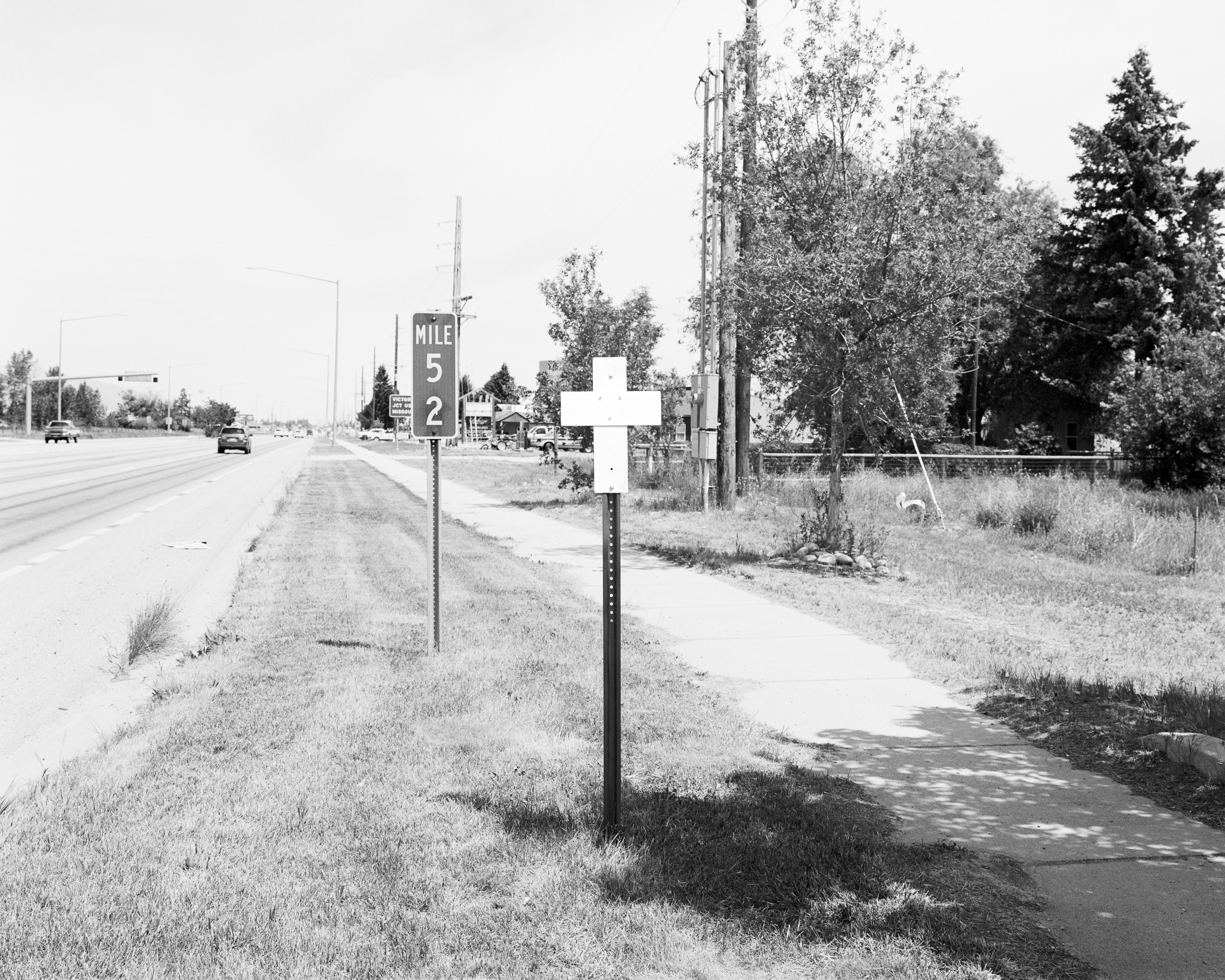 Click to Enlarge Black-and-white photograph of white cross on a pole between sidewalk and highway, adjacent to a sign reading MILE 52.
