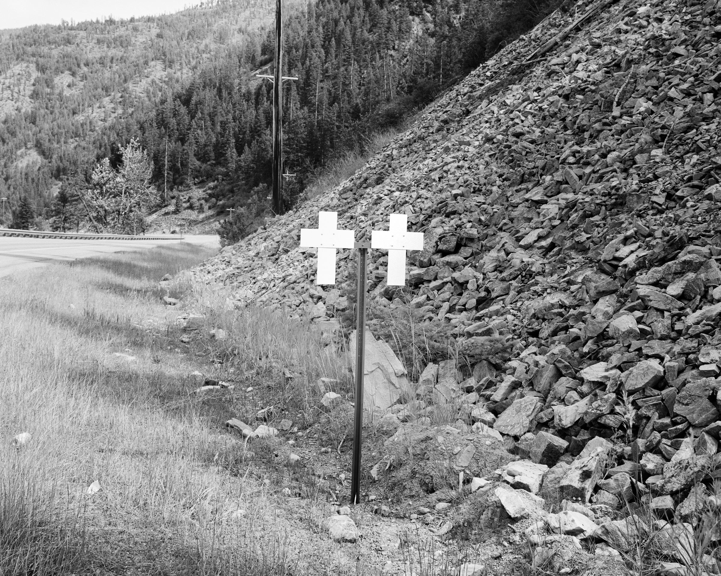 Click to Enlarge Black-and-white photograph of two white crosses on a pole between rocky hillside and roadway.