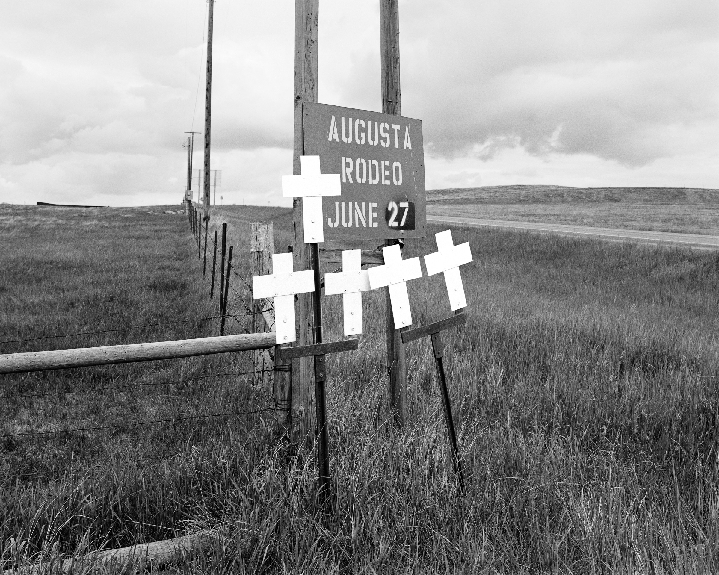 Click to Enlarge Black-and-white photograph of three white crosses on one pole and two on another, in a grassy field beside sign reading AUGUSTA RODEO JUNE 27.