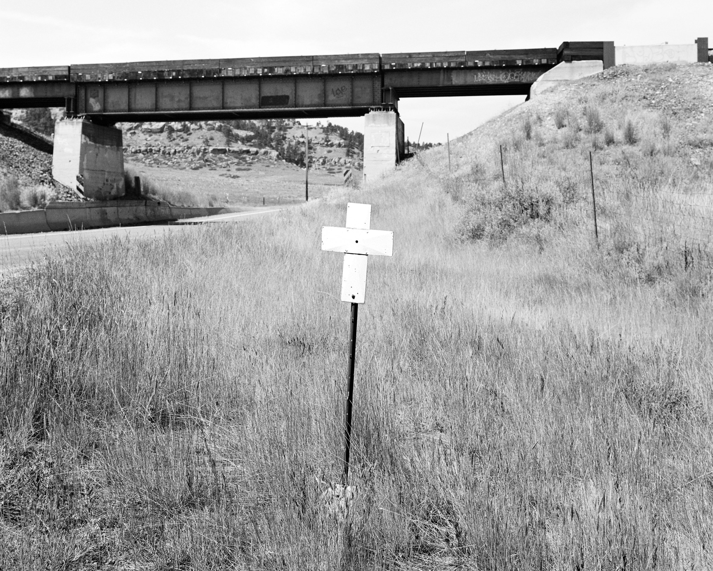Click to Enlarge Black-and-white photograph of white cross on a pole beside rural roadway, with overpass in midground.