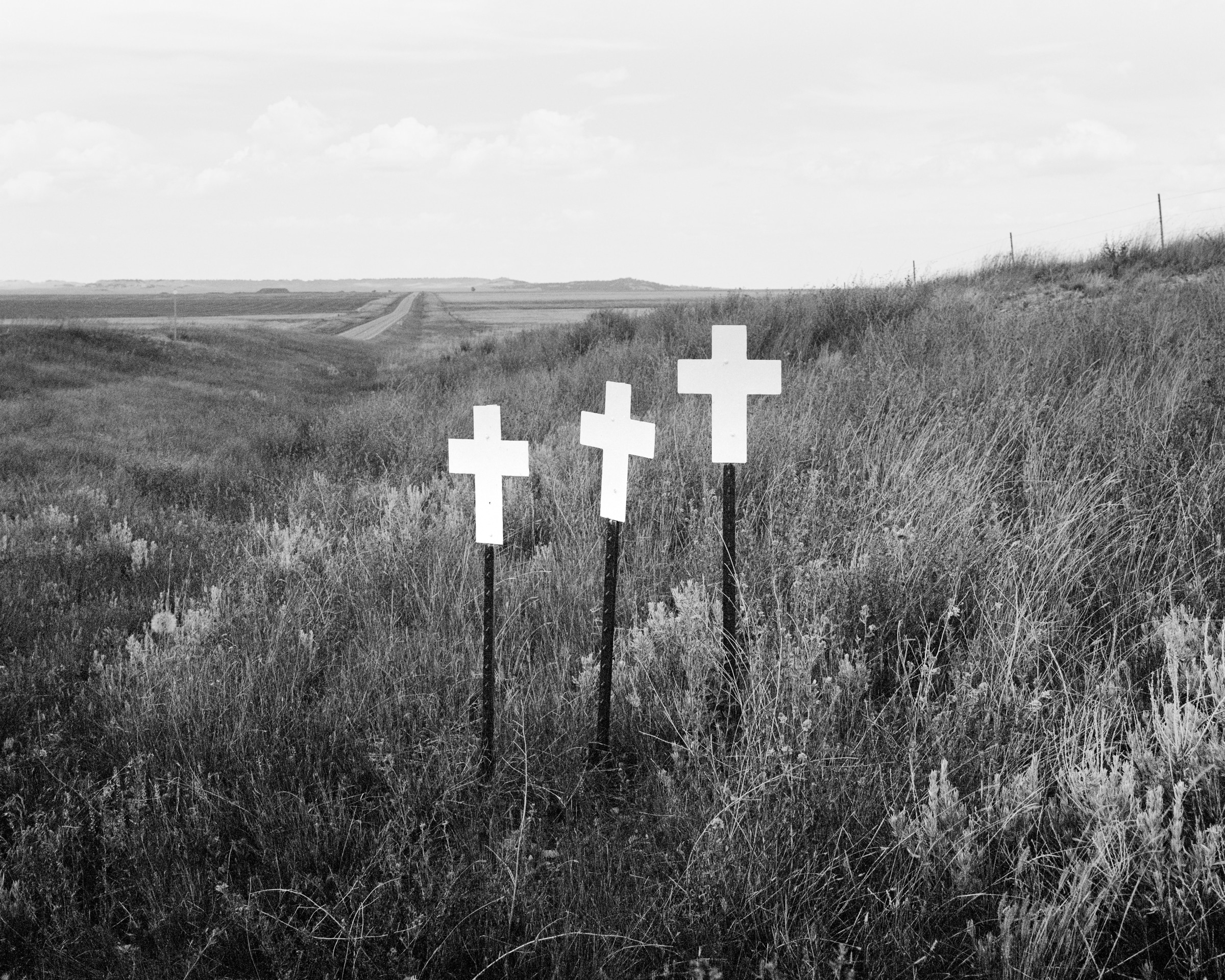 Click to Enlarge Black-and-white photograph of three white crosses, each on a pole in grassy field with road in the distance.