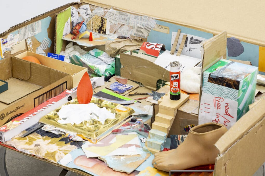 A close-up view of an artwork. We see a cardboard box with many common objects and other bits of cardboard arranged in a messy and chaotic that resembles the floorplan of a home. One of the largest items is a hollow plastic foot.
