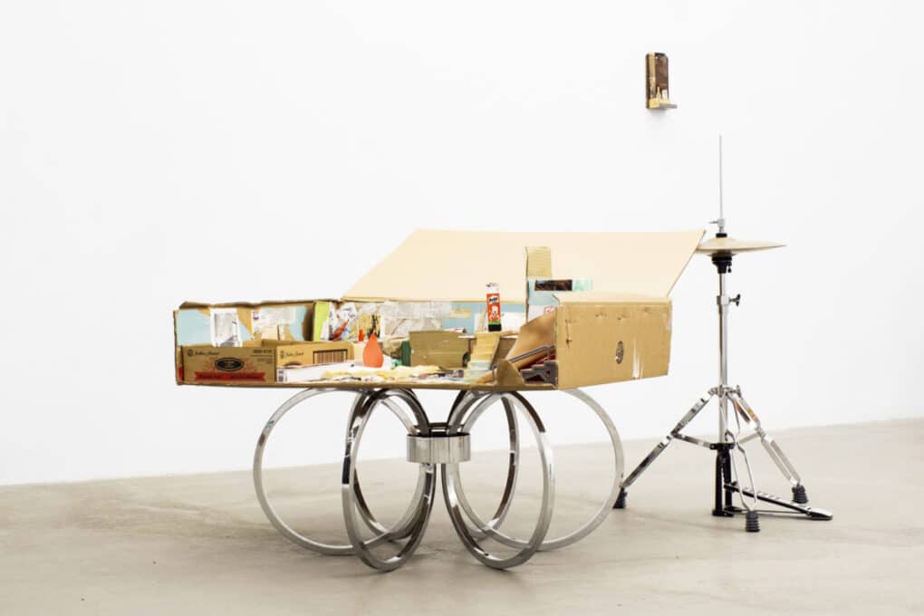 A sculpture in a gallery setting shows an open cardboard boxes, with various things inside, on a base made from silver rings. Slightly behind and to the side is the cymbal from a drum kit.