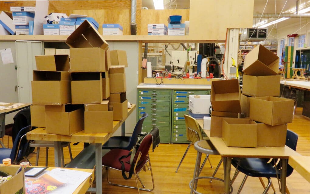 Two desks in an empty but visually busy classroom are each stacked with a dozen or so cardboard boxes in haphazard fashion.