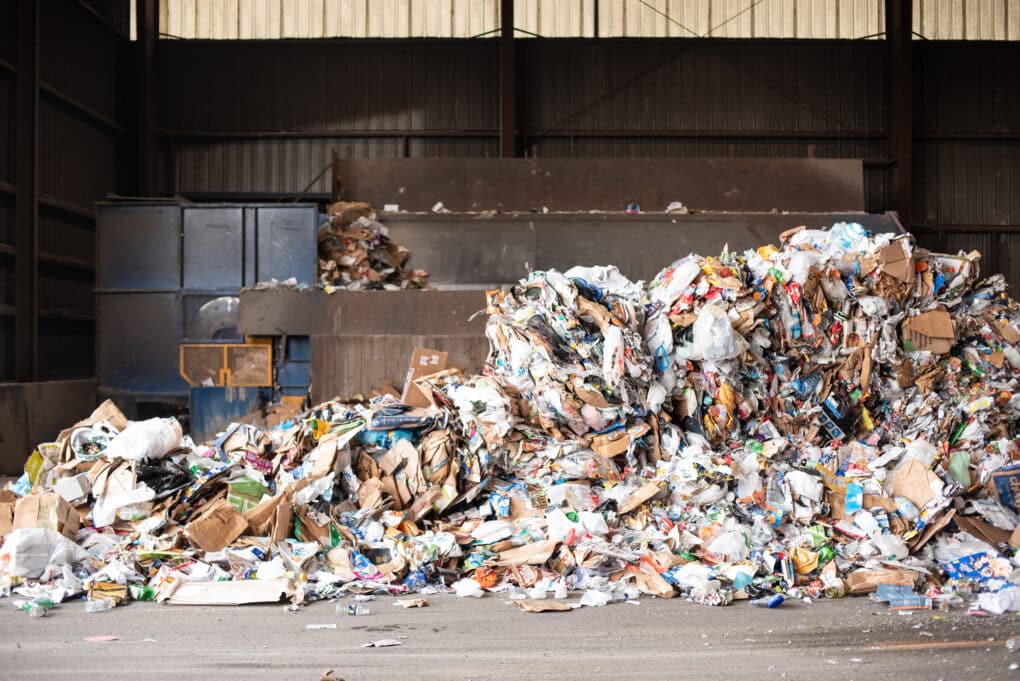 A large pile of mixed household waste, including a lot of cardboard, is shown in a warehouse, apparently a waste transfer facility.